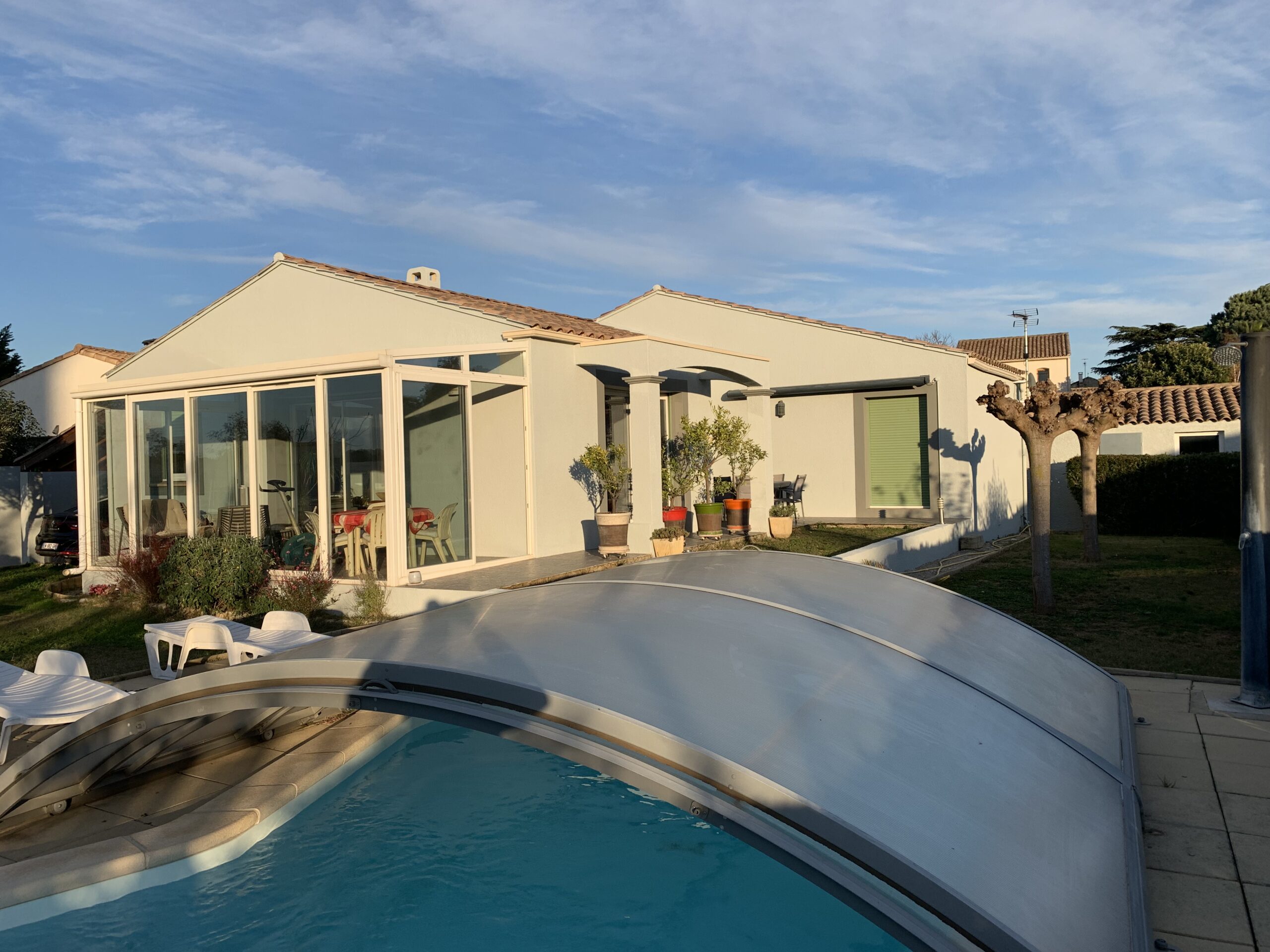 Une maison blanche moderne avec de grandes fenêtres en verre, traitées avec une peinture extérieure durable, se trouve à côté d'une piscine couverte d'un abri transparent ; le soleil projette de longues ombres sur le jardin et le mobilier de terrasse.