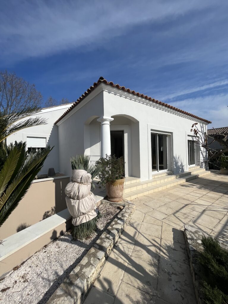 Une maison blanche moderne avec un toit en tuiles rouges, des colonnes à l'entrée et de grandes fenêtres témoigne d'une rénovation récente. Des plantes en pot, une allée pavée, une sculpture de jardin et de la verdure luxuriante agrémentent la cour d'entrée sous un ciel bleu.