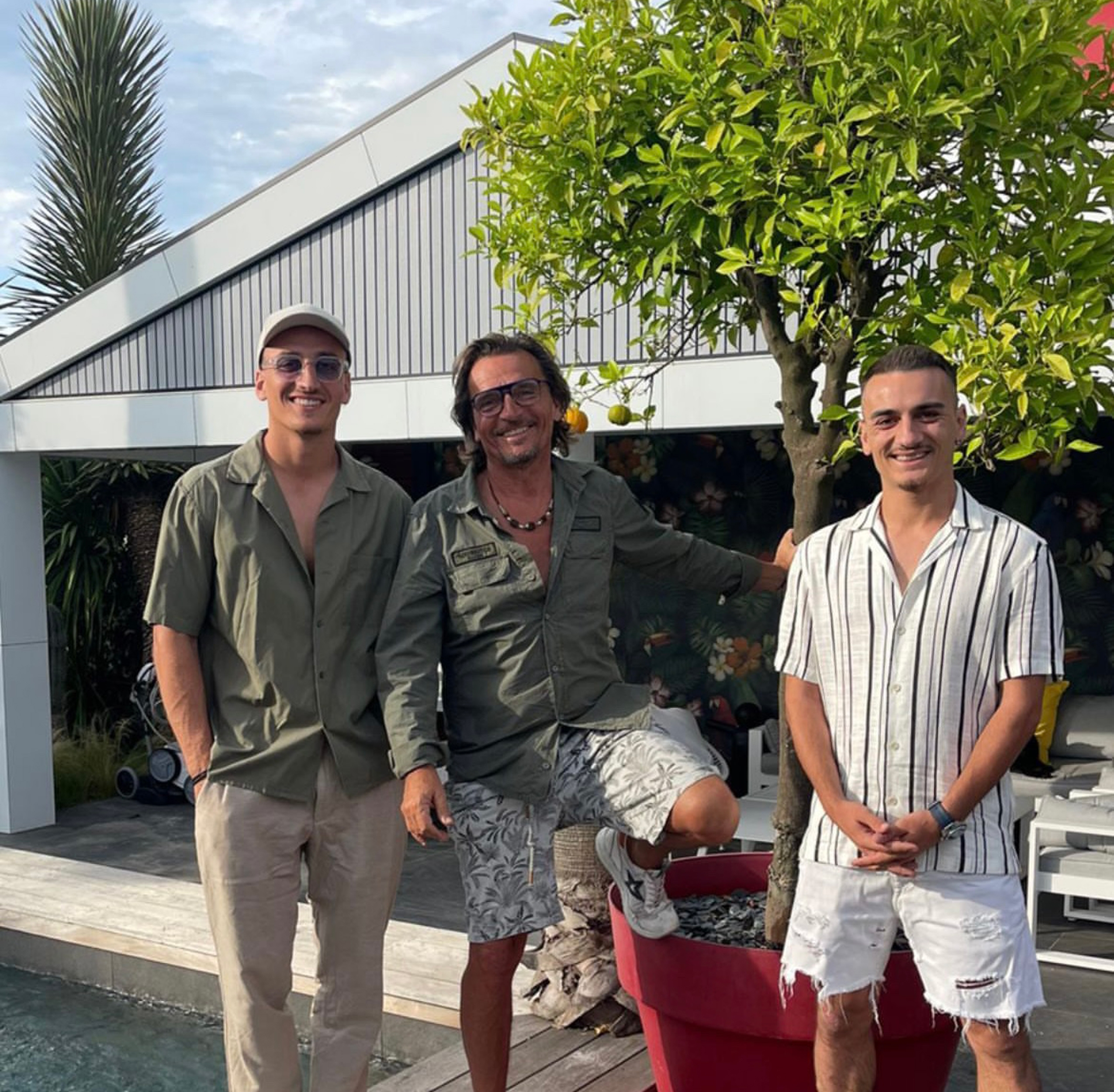 Trois hommes sourient et posent en plein air au bord d'une piscine. L'un d'eux utilise une technique ludique, en posant le pied sur un grand pot de fleurs rouge tenant un petit arbre. Les autres se tiennent à ses côtés, avec un bâtiment moderne en arrière-plan.