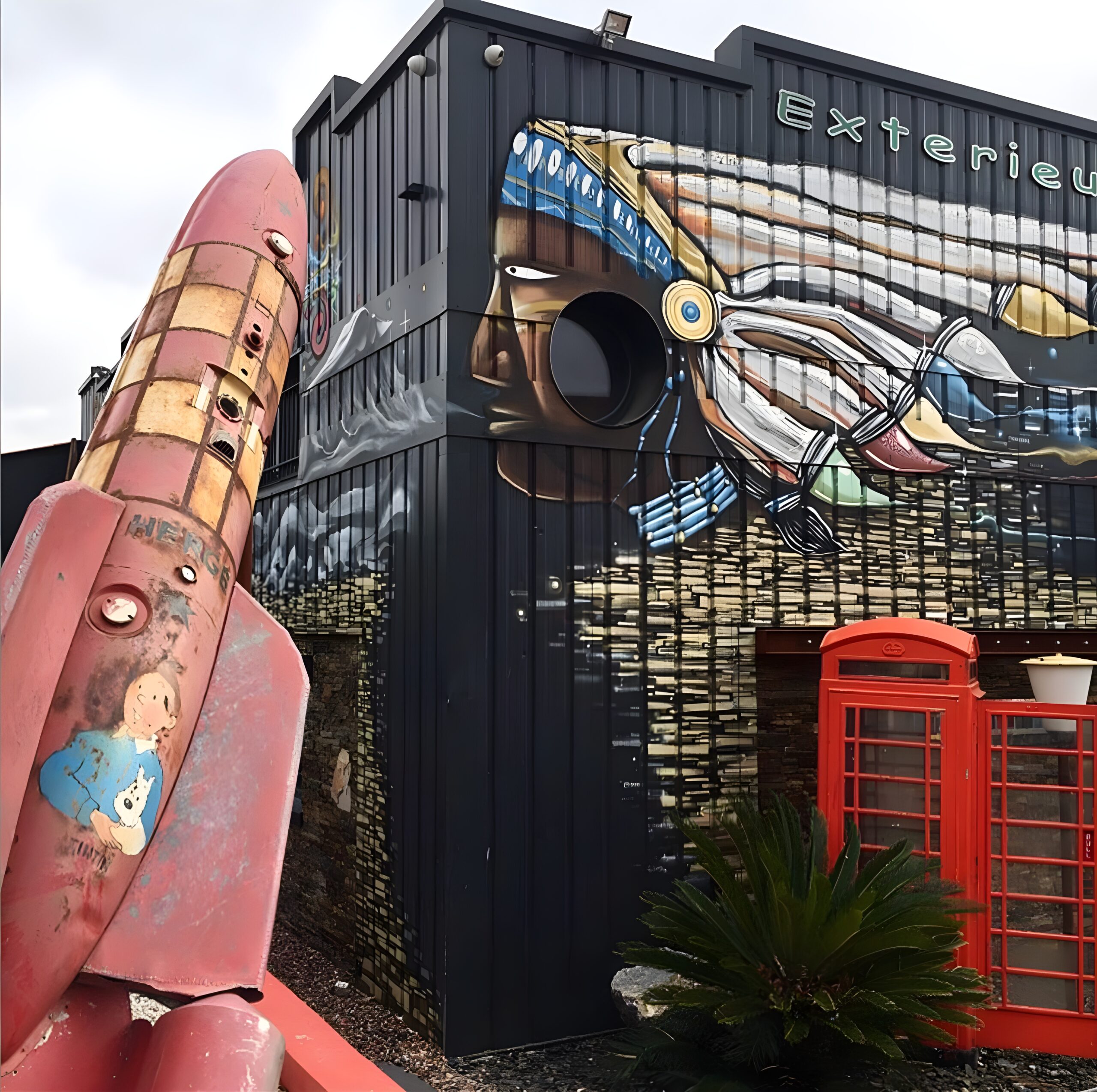 Une fusée rouge à l'effigie d'un personnage de dessin animé se trouve devant un bâtiment noir orné d'une grande peinture murale représentant un calmar et d'une cabine téléphonique rouge classique. La scène est colorée et fantaisiste.