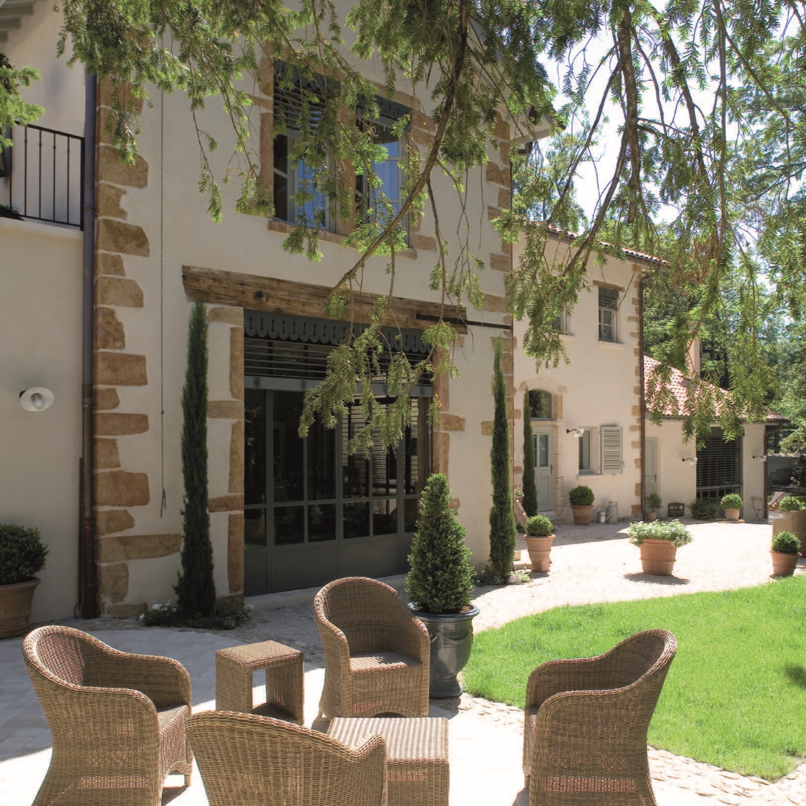 Dans une cour ensoleillée, des chaises en osier et une table sont disposées sur un patio en pierre, entouré de plantes en pot et de buissons bien entretenus. L'arrière-plan montre une charmante maison aux accents de pierre et aux grandes fenêtres, entourée de verdure.