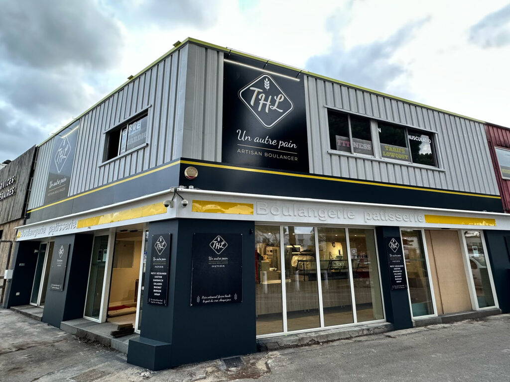 La devanture d'une boulangerie moderne nommée Un autre pain, avec de grandes fenêtres, des garnitures noires et jaunes et une enseigne indiquant Boulangerie Pâtisserie. L'intérieur est visible à travers les fenêtres.